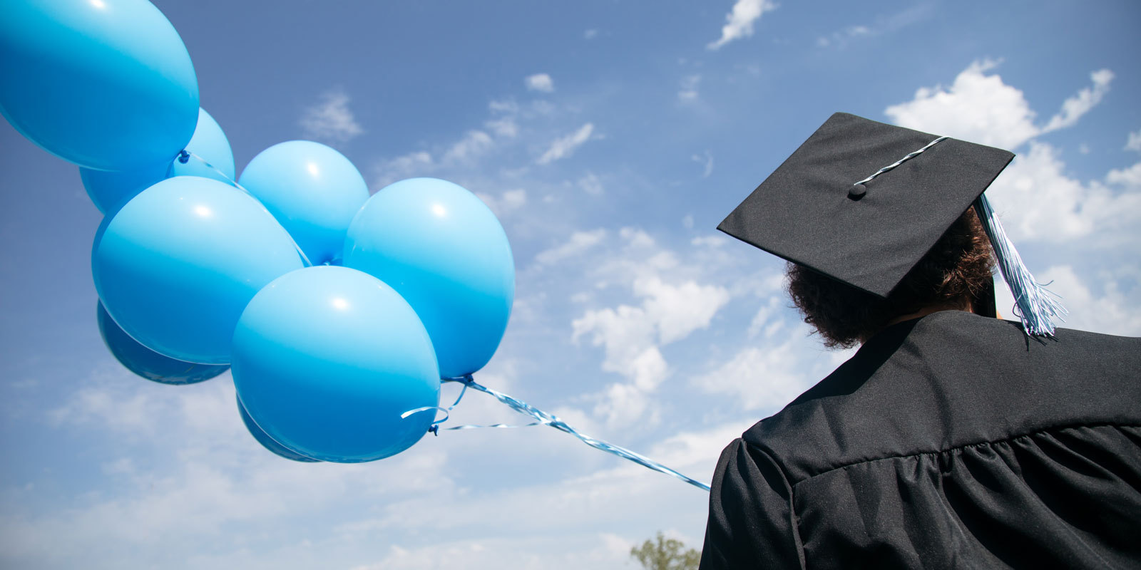 Graduate in cap and gown stands outside with blue balloons